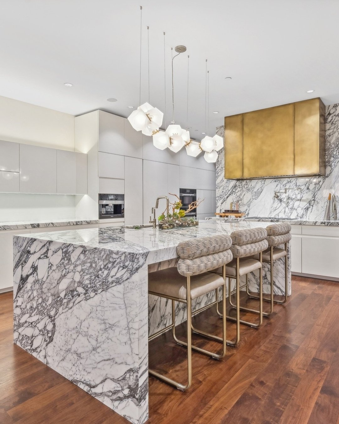 Cluster of geometric white glass pendants hanging over a marble waterfall island with gold accents.