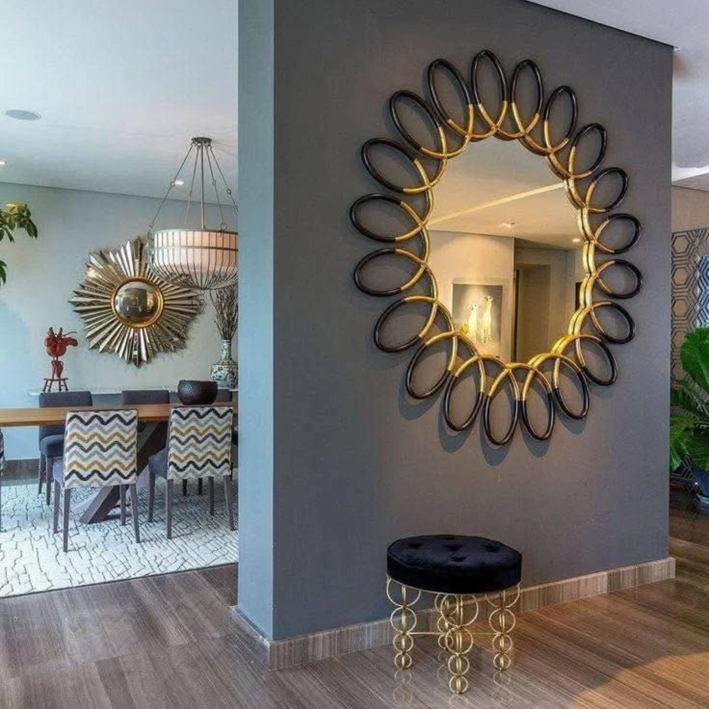 Large round mirror with black and gold chain-link frame mounted on a dark grey partition wall above a black velvet stool.