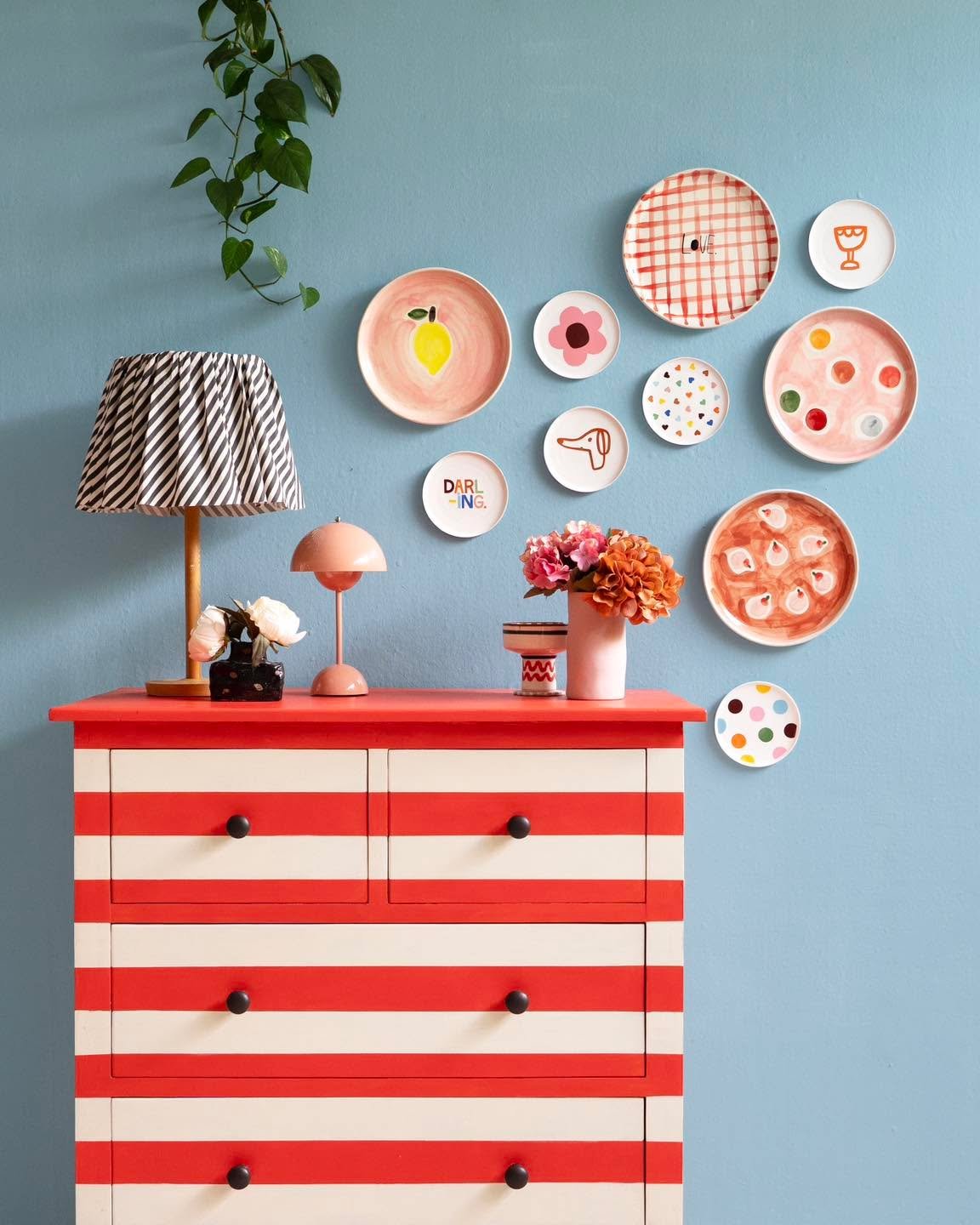 Blue wall decorated with colorful whimsical plates above a red and white striped chest of drawers