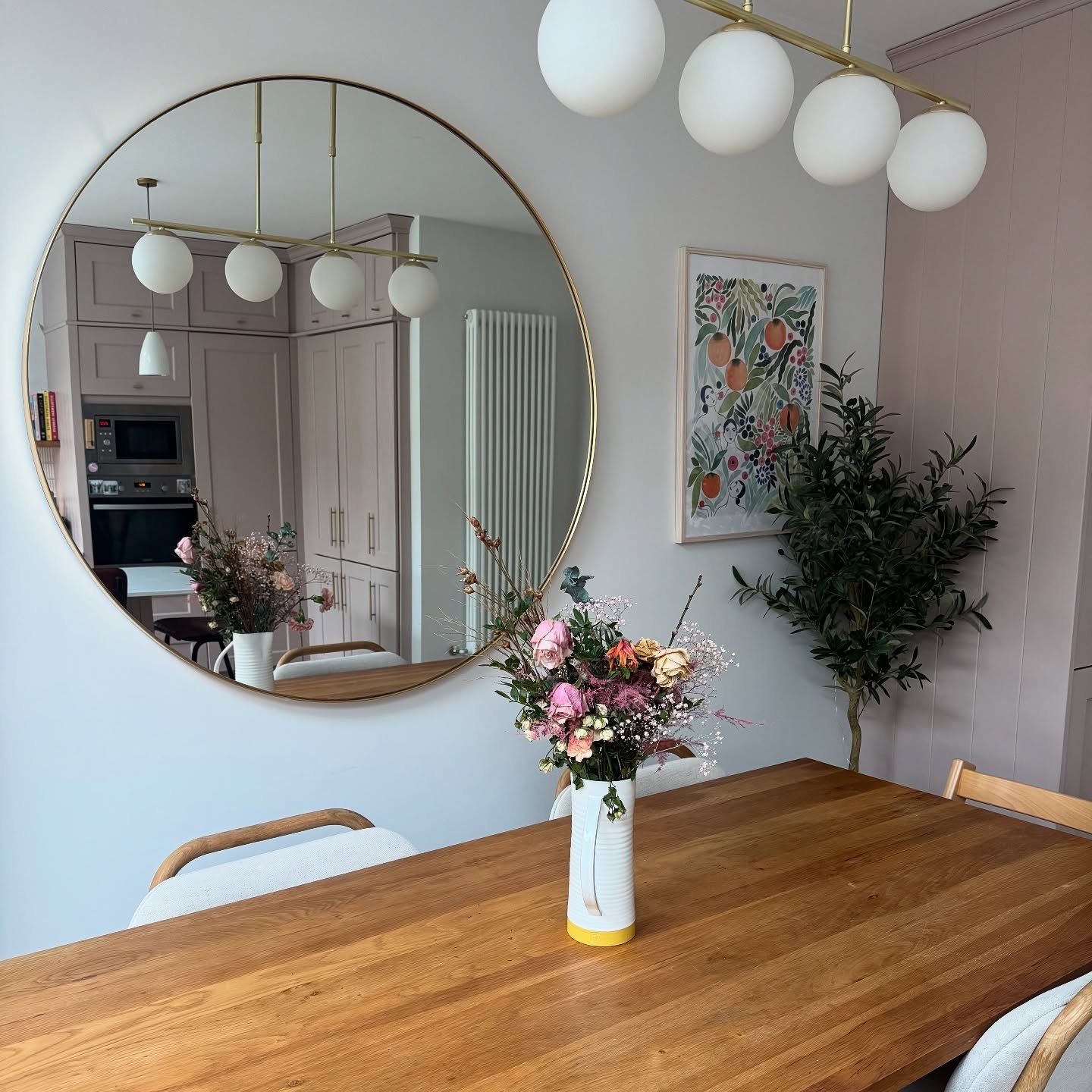 Large circular gold mirror hanging in a dining room reflecting a pink kitchen and globe lights.