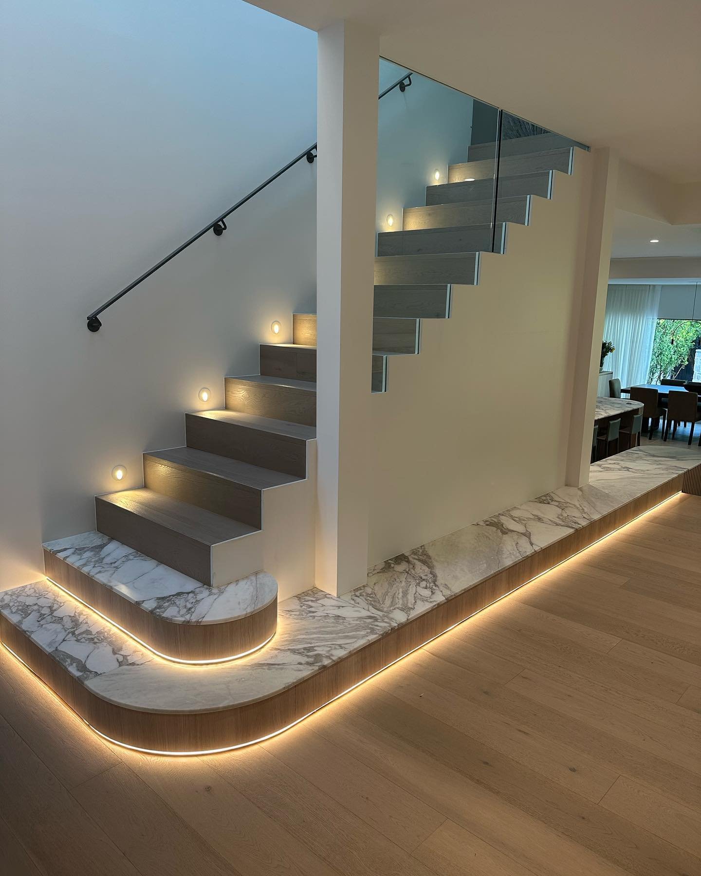 Modern staircase featuring a curved marble base platform with under-cabinet warm LED lighting and recessed wall step lights.