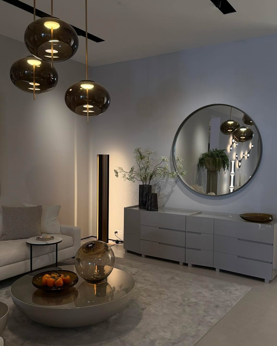 Modern living room featuring a grey accent wall with a large round mirror, illuminated by a corner floor lamp and hanging smoked glass pendants.