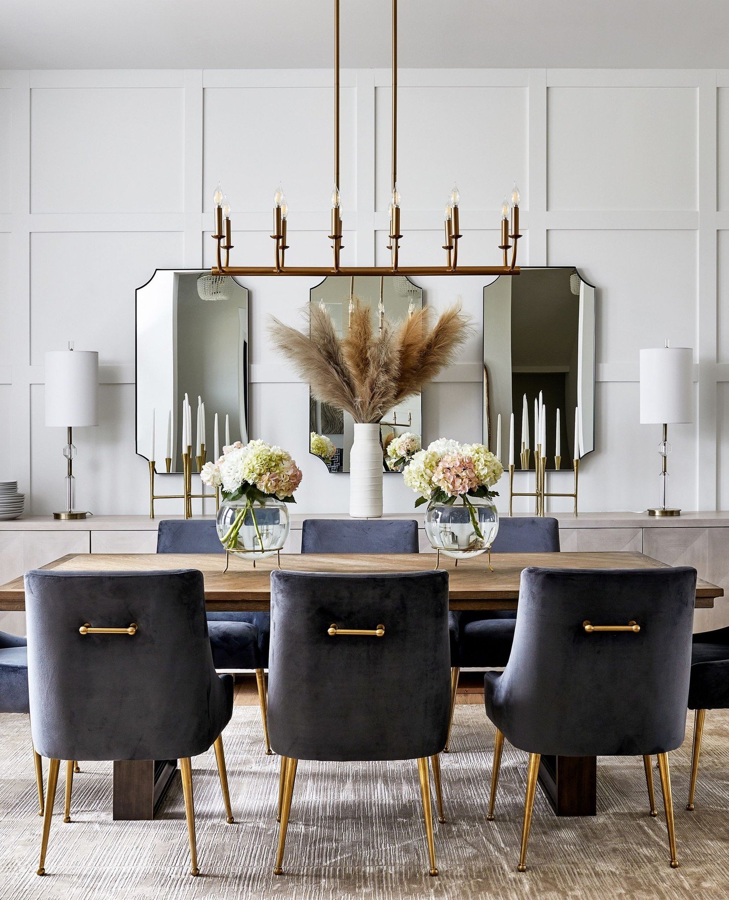 Linear gold candle chandelier hanging over a wood dining table with blue velvet chairs and three wall mirrors.
