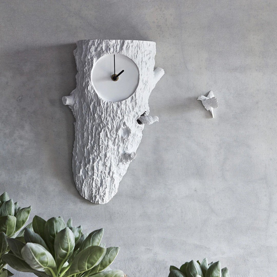 White tree trunk shaped wall clock with small birds mounted on a grey concrete wall